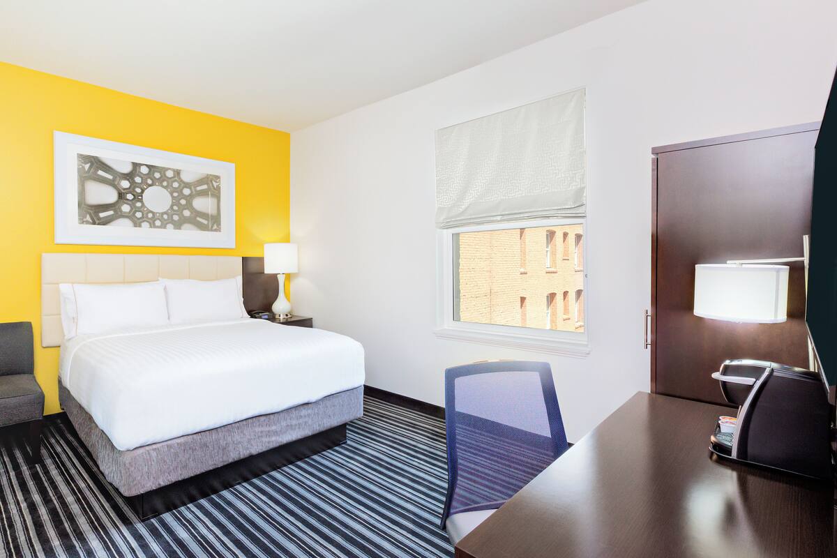 A hotel room with a bed, a desk, a chair, a lamp, and a window with a view of buildings.