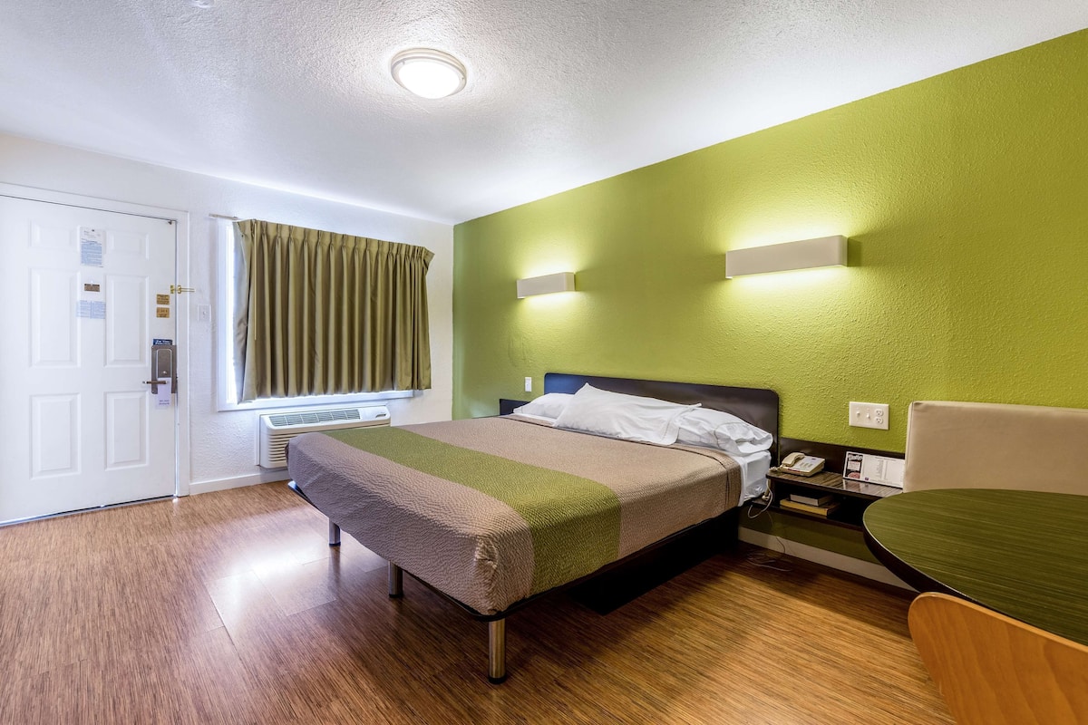 A hotel room with a bed, a bedside table, a chair, a window with curtains, a door, and a wall-mounted light fixture.