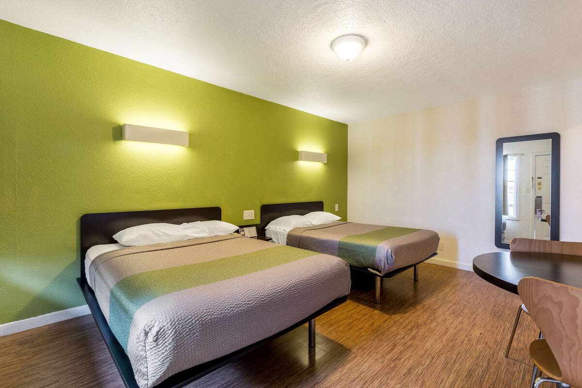 A hotel room with two beds, a wooden floor, a green wall, and a small table with a chair.
