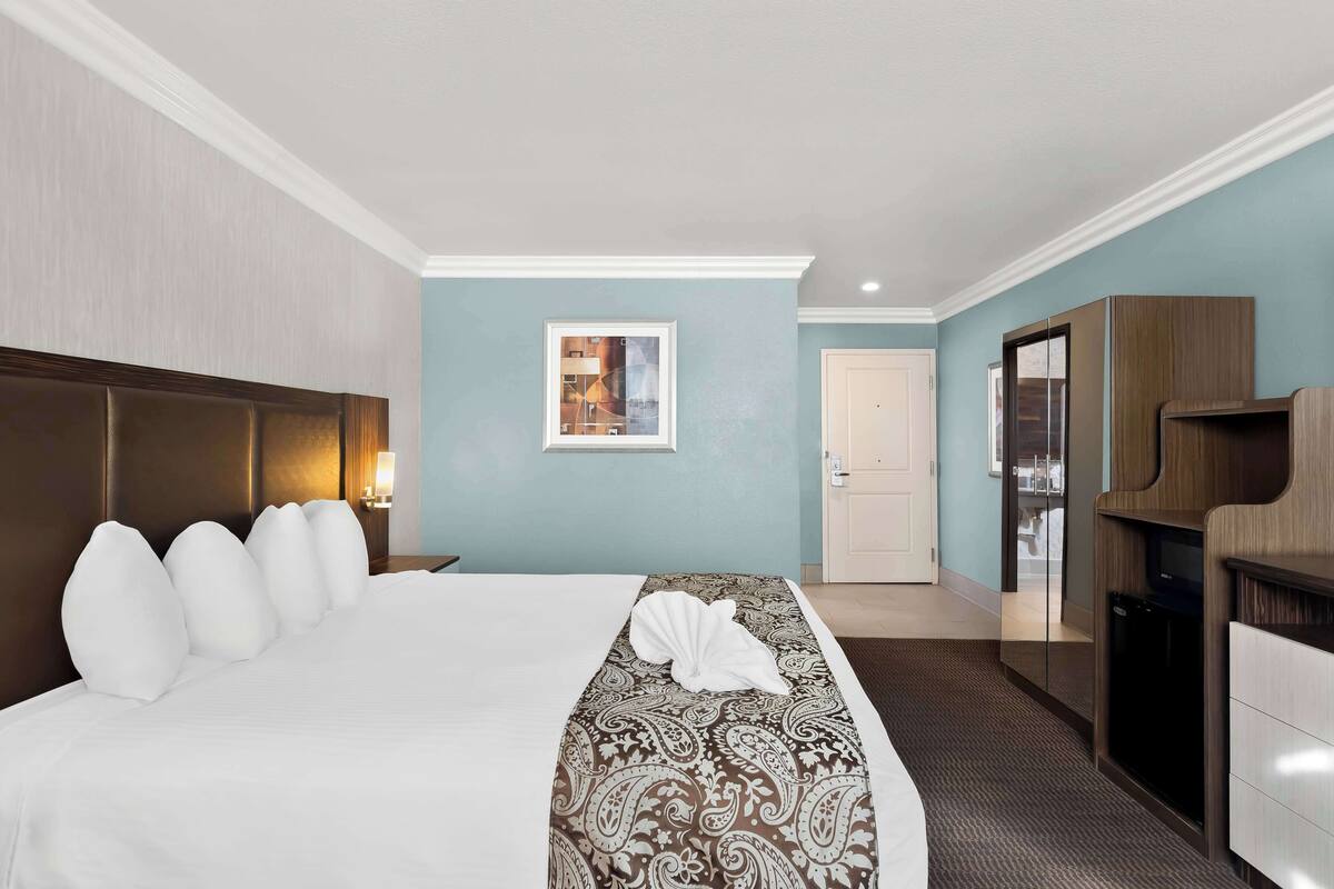 A hotel room with a large bed, a wooden headboard, a bedside table with a lamp, a wardrobe, and a door leading to a bathroom.