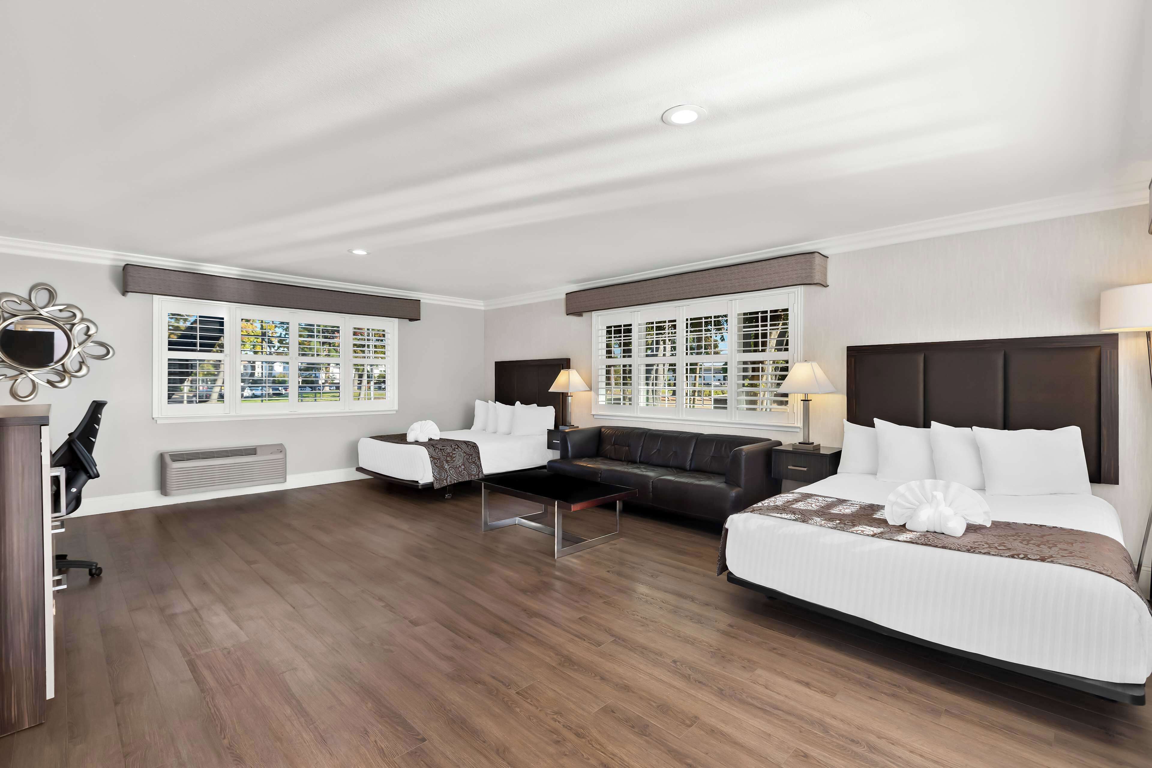 A hotel room with a large bed, a sofa, a desk, and a chair. There are two windows with blinds, a ceiling light, and a wall-mounted mirror.