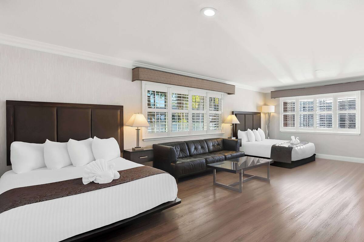 A hotel room with a large bed, a sofa, a coffee table, and two windows with blinds.