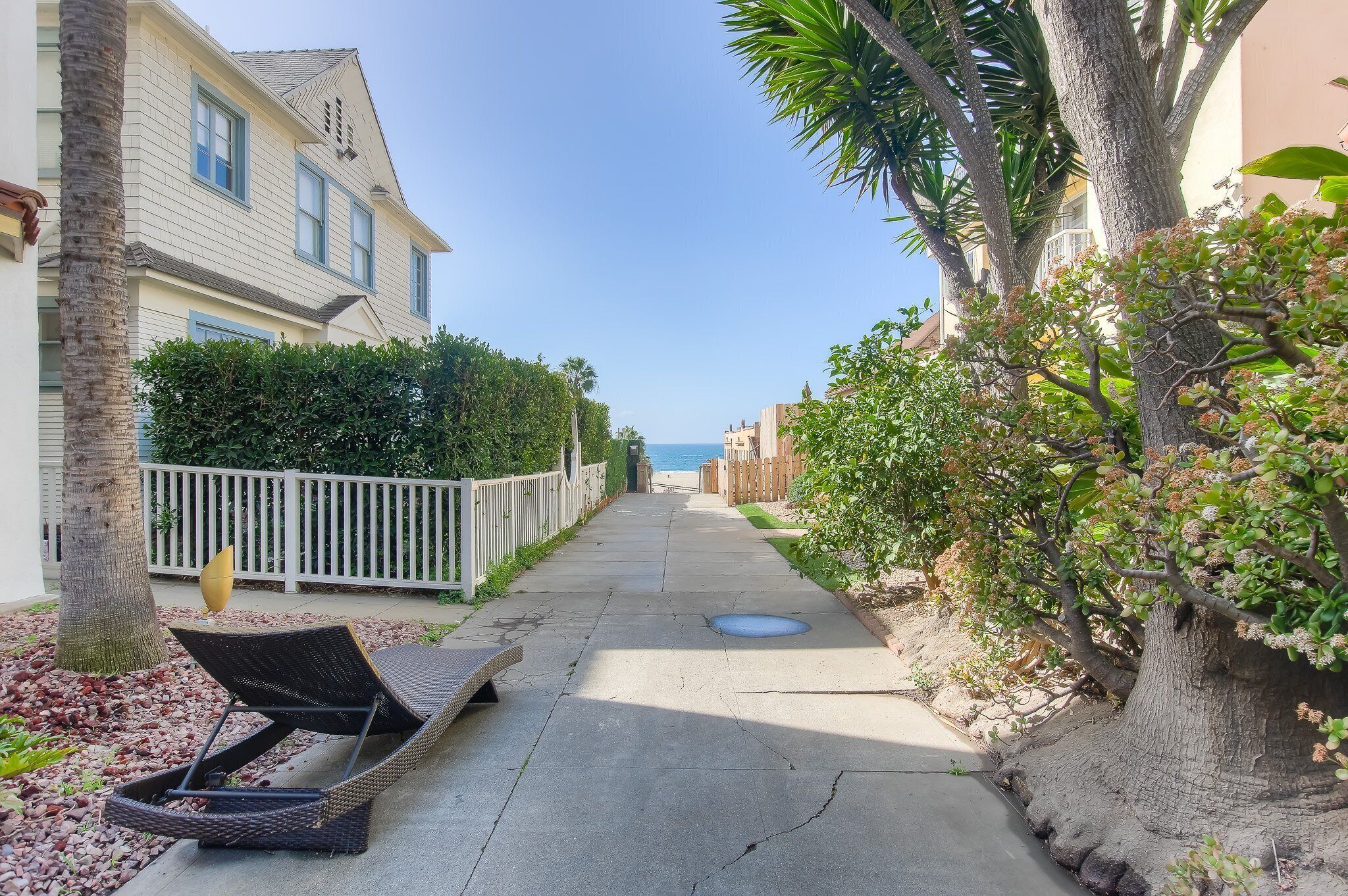 A narrow pathway flanked by houses and trees, leading to a beach.
