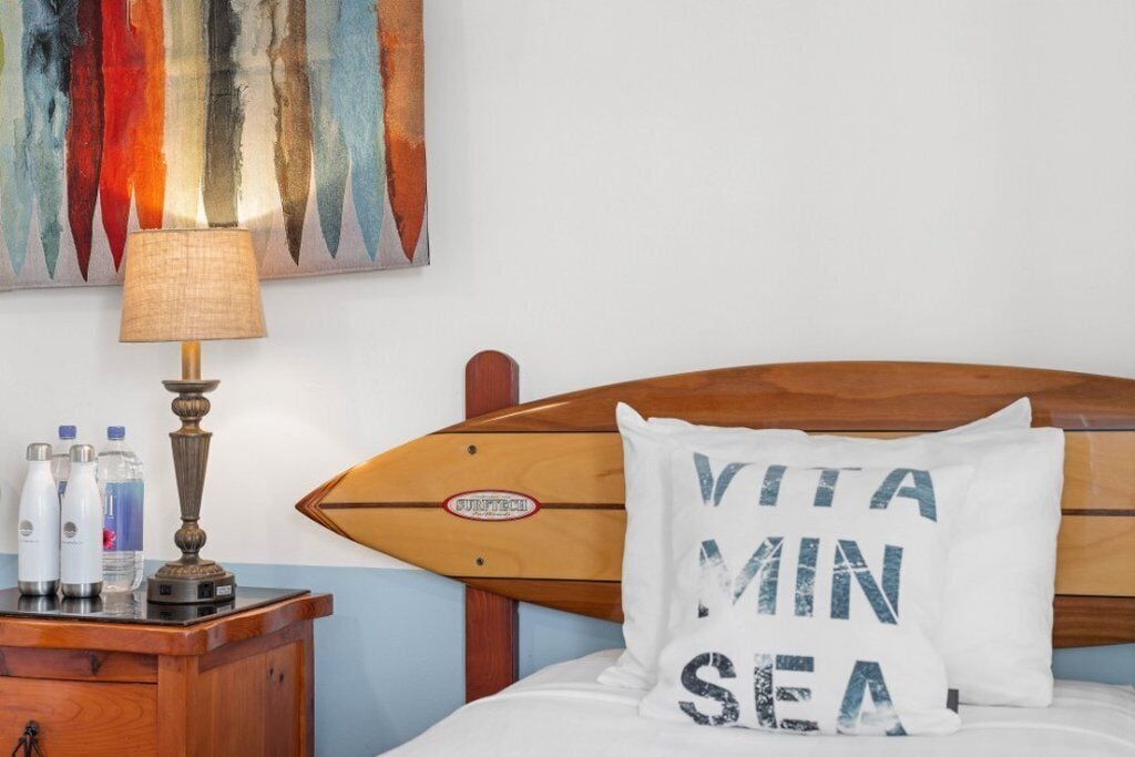 Uma cama com cabeceira em formato de prancha de surf, um travesseiro branco com o texto "VITA MIN SEA", uma luminária e uma mesa de cabeceira de madeira com garrafas de água.