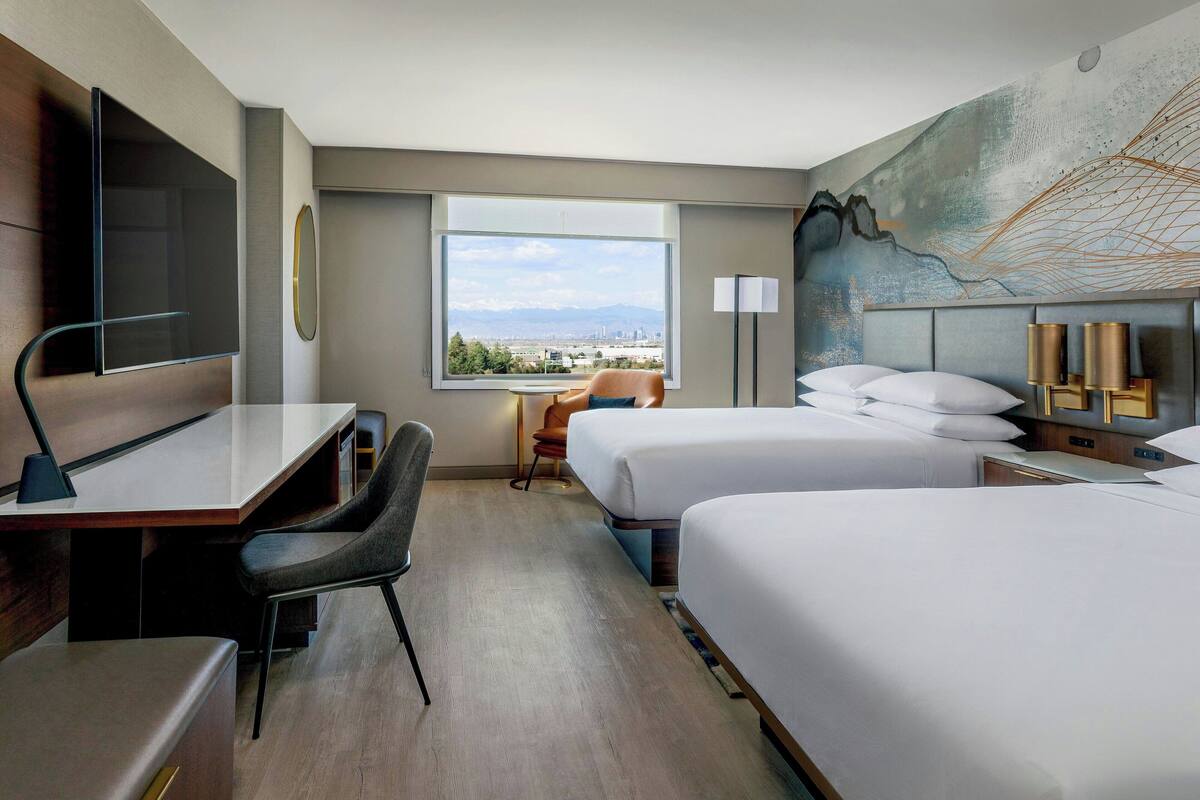 Ett hotellrum med ett skrivbord, två sängar, en stol, en TV och ett stort fönster med utsikt över staden.
