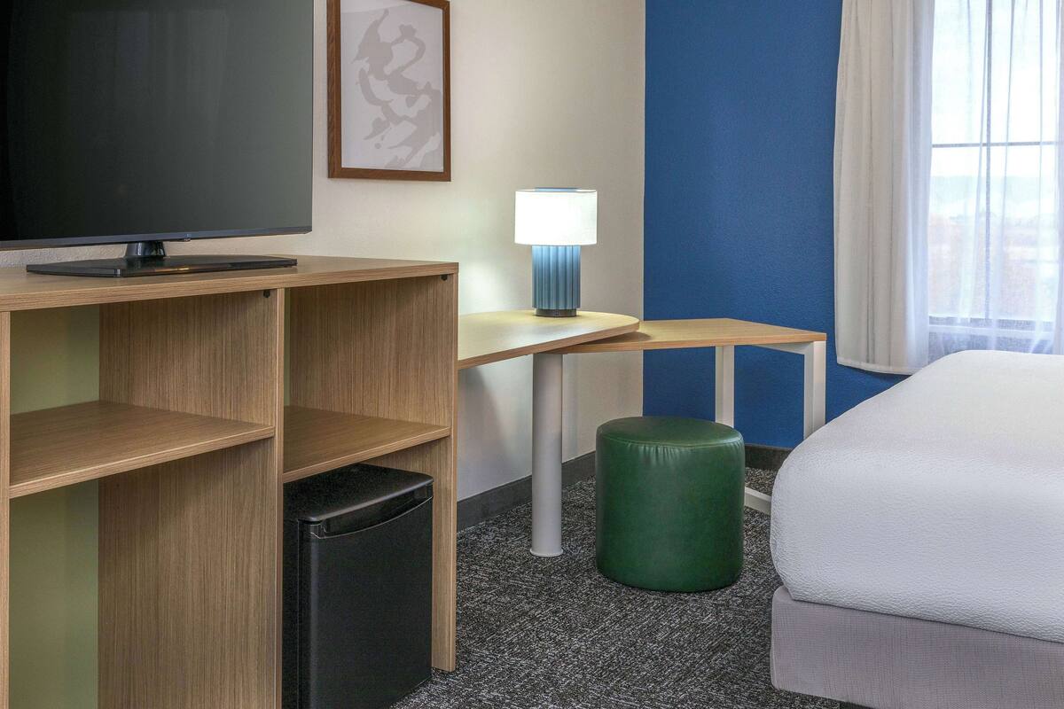 Ein Hotelzimmer mit Flachbildfernseher, Schreibtisch mit Lampe, Pouf in Grün, Bett und Fenster mit Vorhängen.