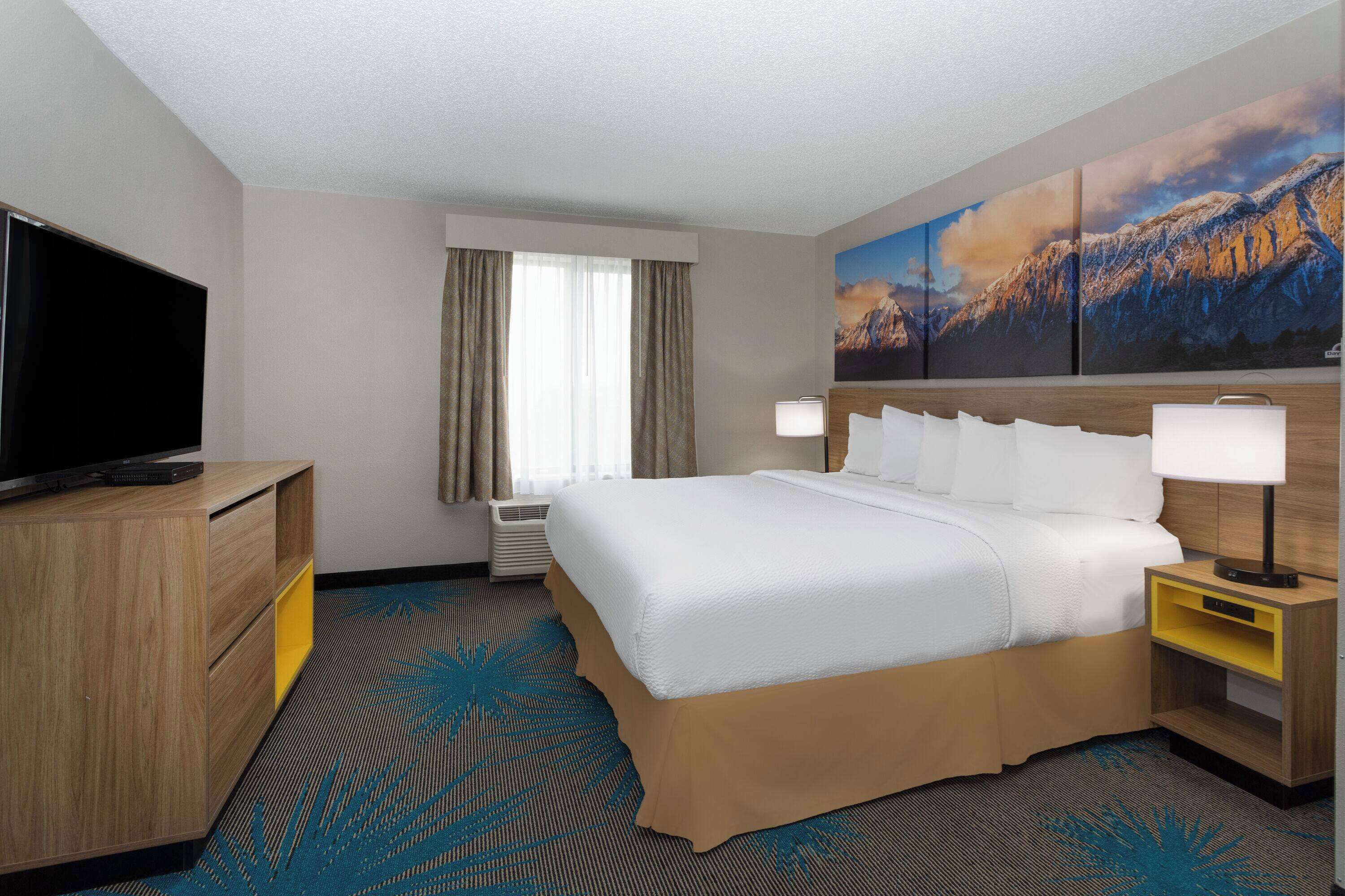 A hotel room with a large bed, a TV, a desk, and a painting of mountains.