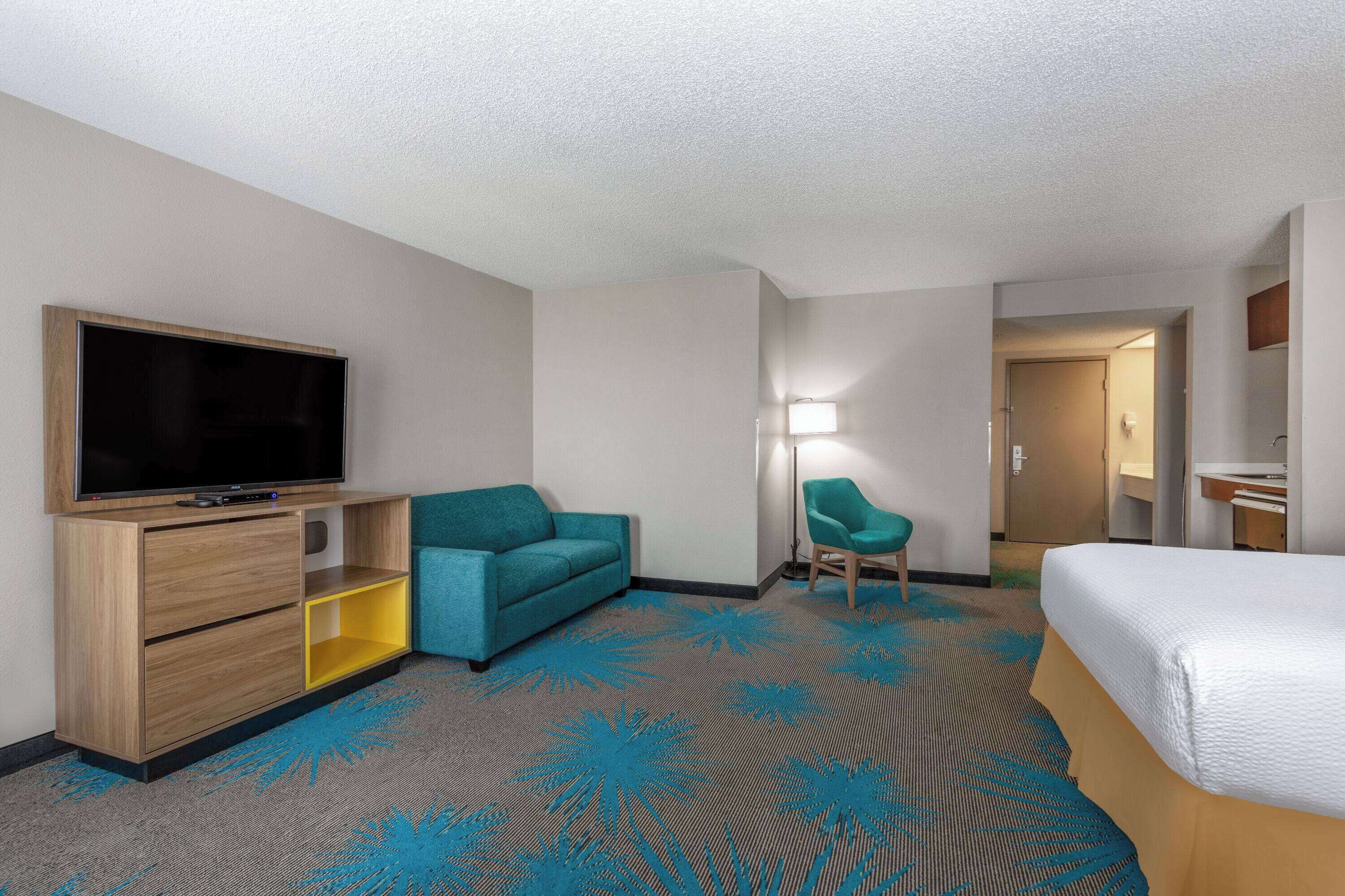 Una camera d'albergo con un letto, una televisione su un mobile in legno, un divano color petrolio, una sedia e un tappeto con un motivo di palme blu.