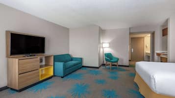 Una camera d'albergo con un letto, una televisione su un mobile in legno, un divano color petrolio, una sedia e un tappeto con un motivo di palme blu.