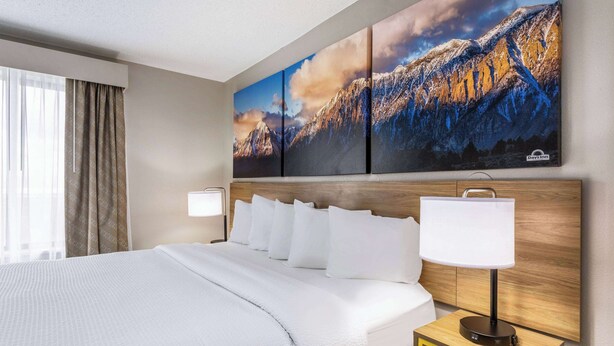 Habitación de hotel con una cama grande, cabecera de madera, mesita de noche con lámpara y un cuadro de un paisaje montañoso en la pared.