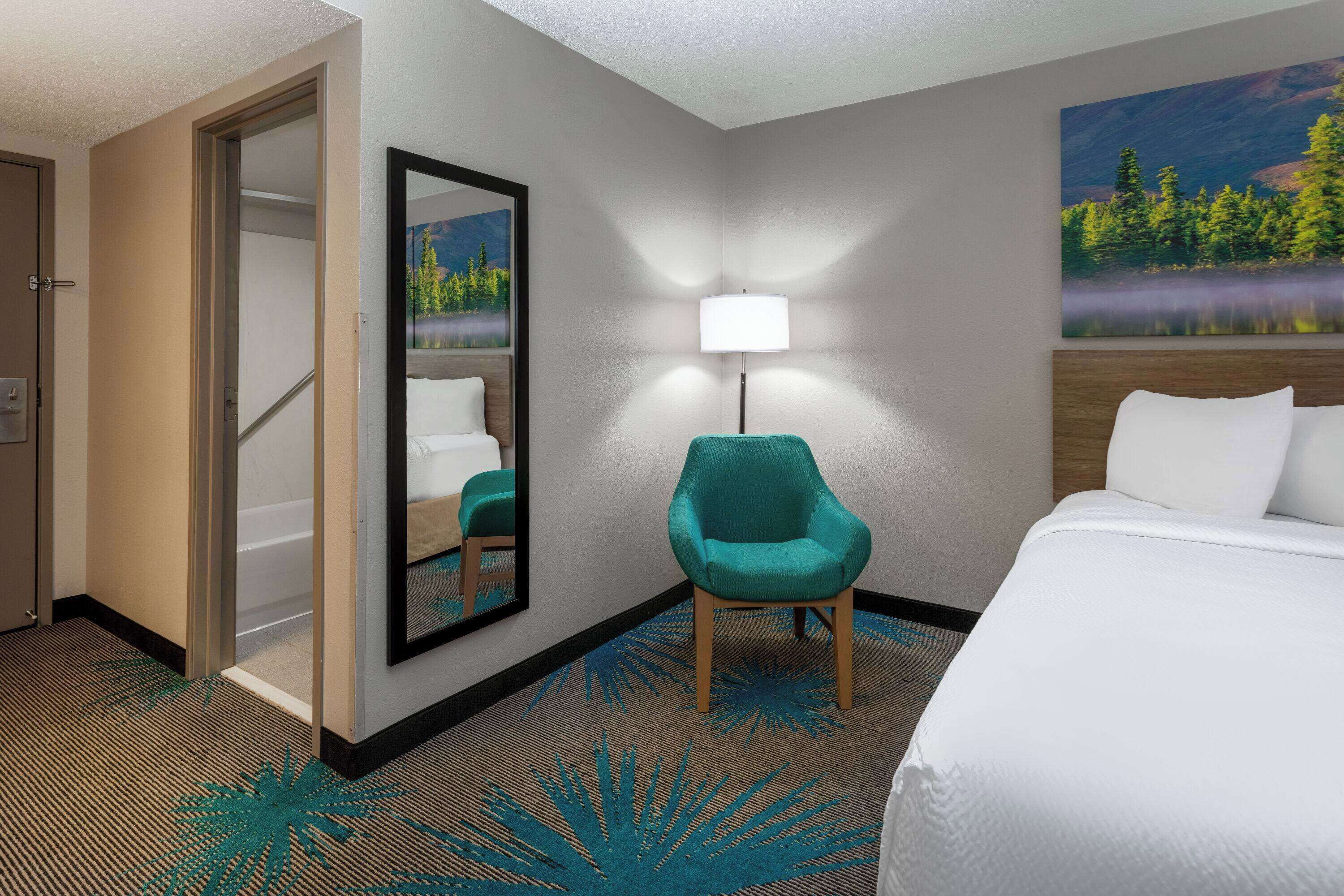 A hotel room with a bed, a teal armchair, a mirror, a lamp, and a painting of a lake.