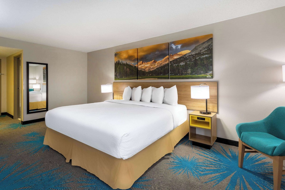 Een hotelkamer met een groot bed, een nachtkastje met een lamp, een stoel en een wanddecoratie van een berglandschap.