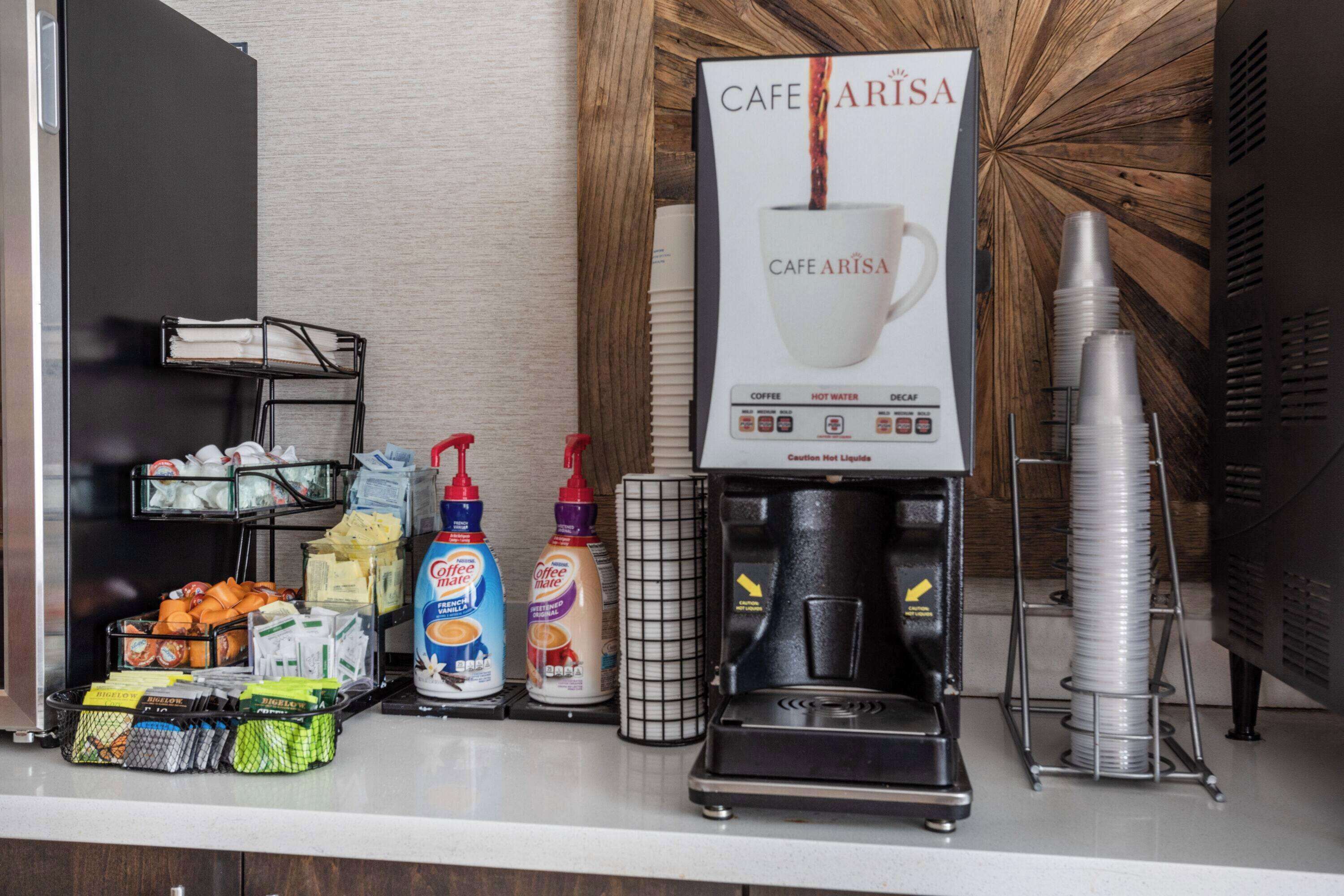 A coffee station with a coffee machine, disposable cups, and various coffee and creamer dispensers.