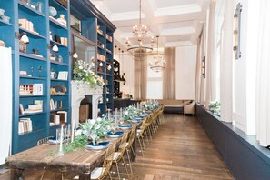 Una larga mesa de comedor con sillas, una estantería y un candelabro en una habitación con suelo de madera y paredes blancas.