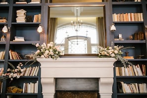 Une bibliothèque avec des étagères, une cheminée et un grand miroir qui reflète un lustre et une fenêtre.