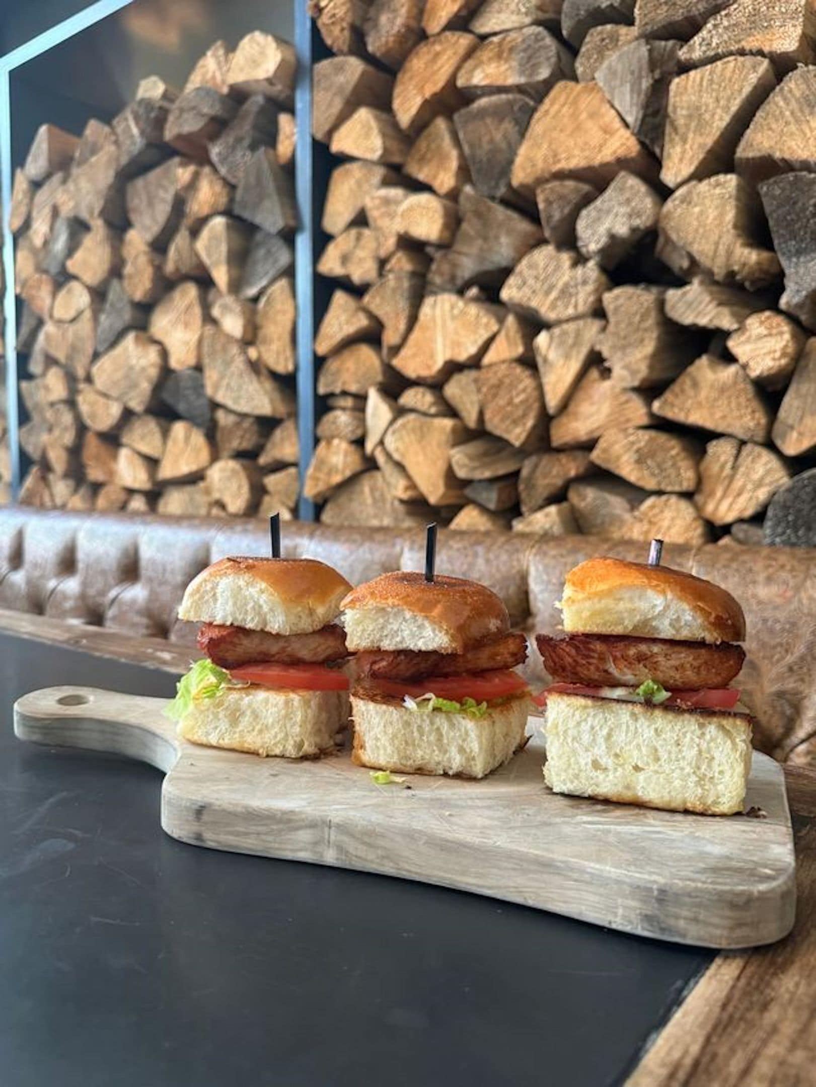 Three sandwiches on a wooden board with a stack of firewood in the background.