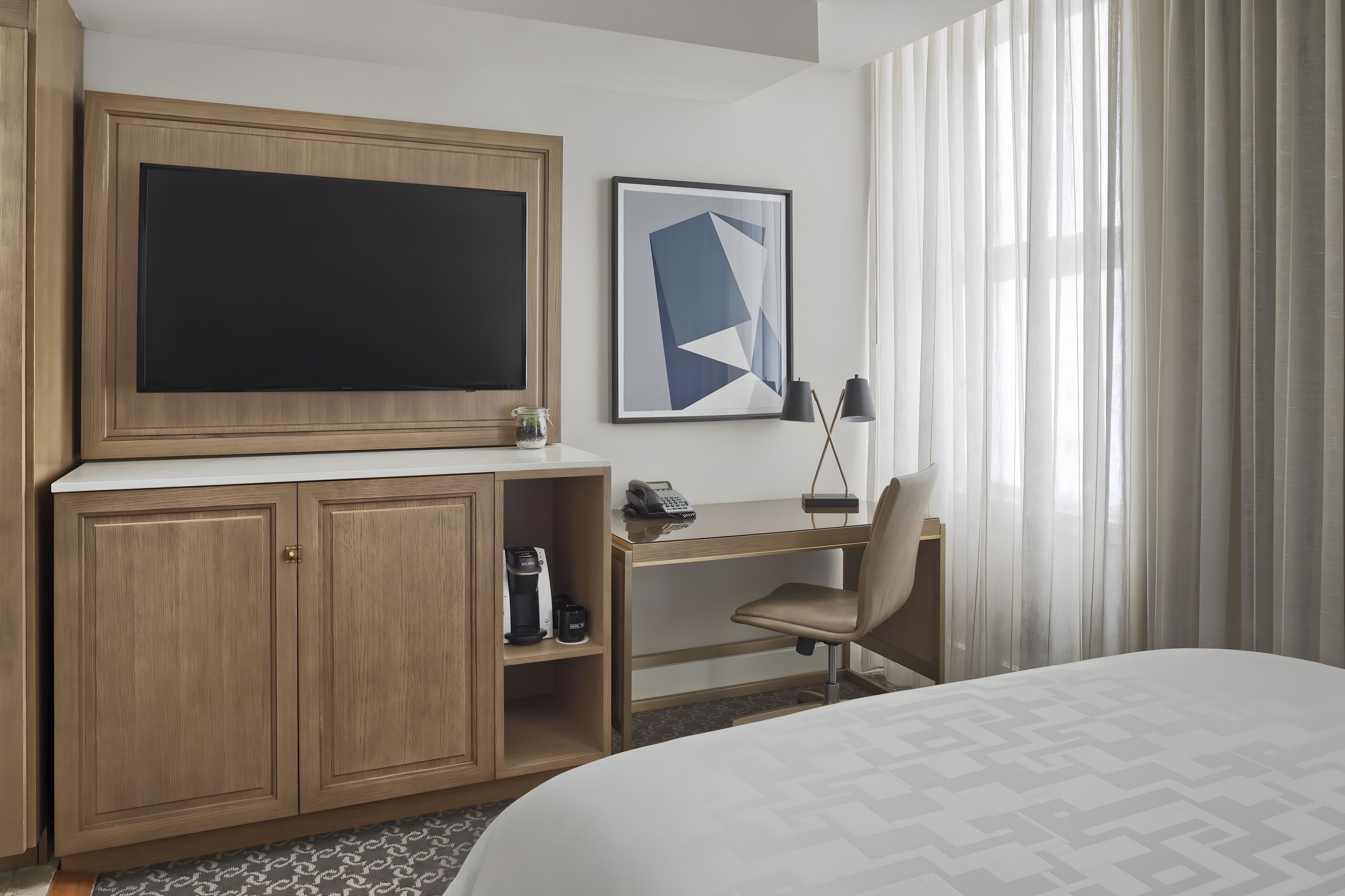 A hotel room with a bed, a desk with a chair, a television, and a framed abstract artwork.