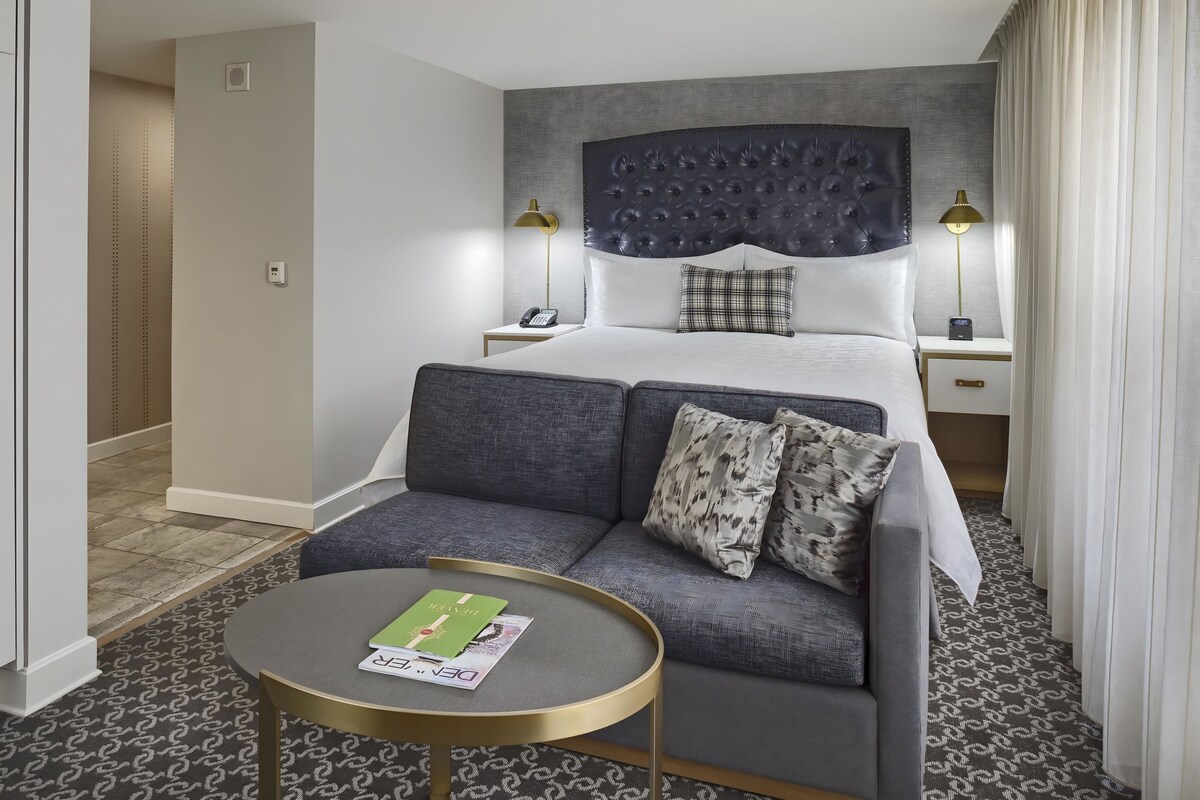 Habitación de hotel con cama, sofá, una mesita, dos lámparas de noche y alfombra estampada.
