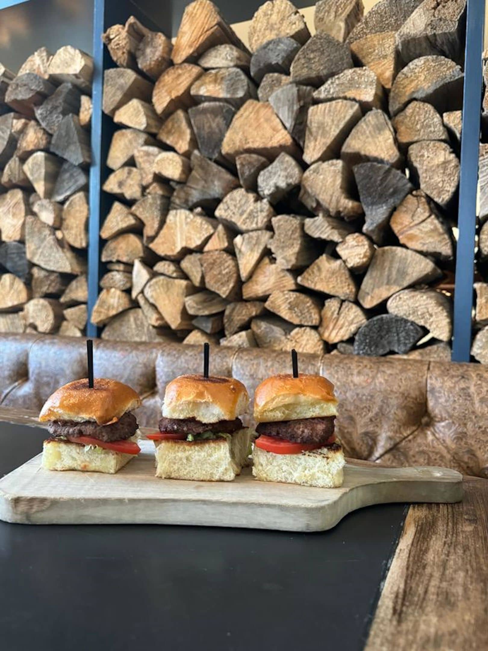 Three burgers on a wooden board with a stack of firewood in the background.