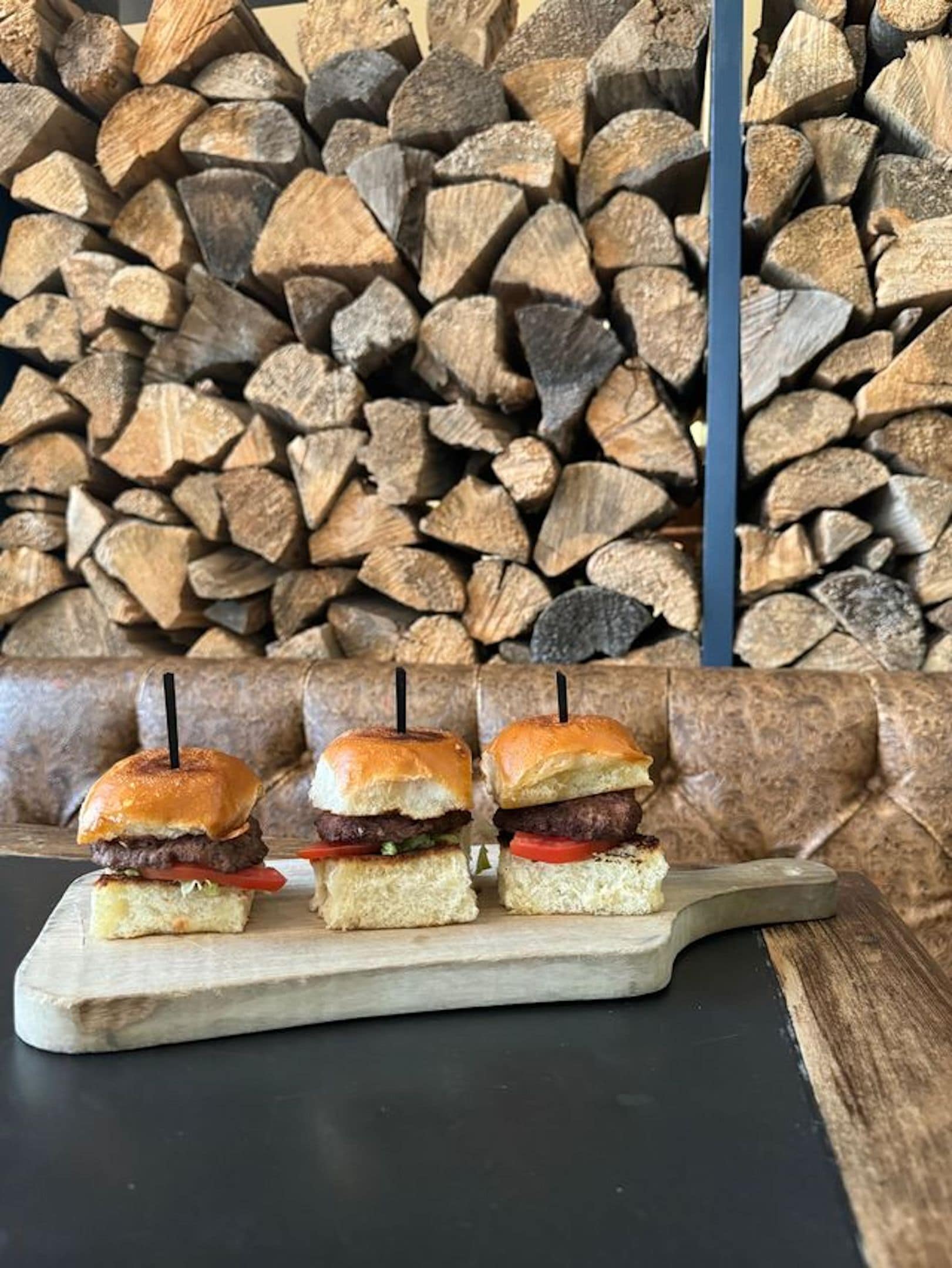 Three burgers on a wooden board with a stack of firewood in the background.