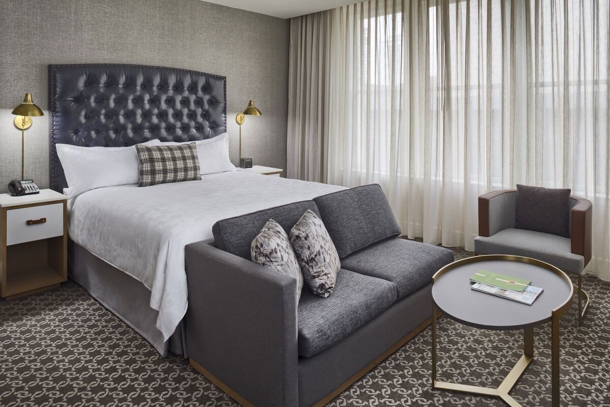 Una habitación de hotel con una cama de cabecera capitonada, un sofá gris, una mesa redonda de centro y una silla.