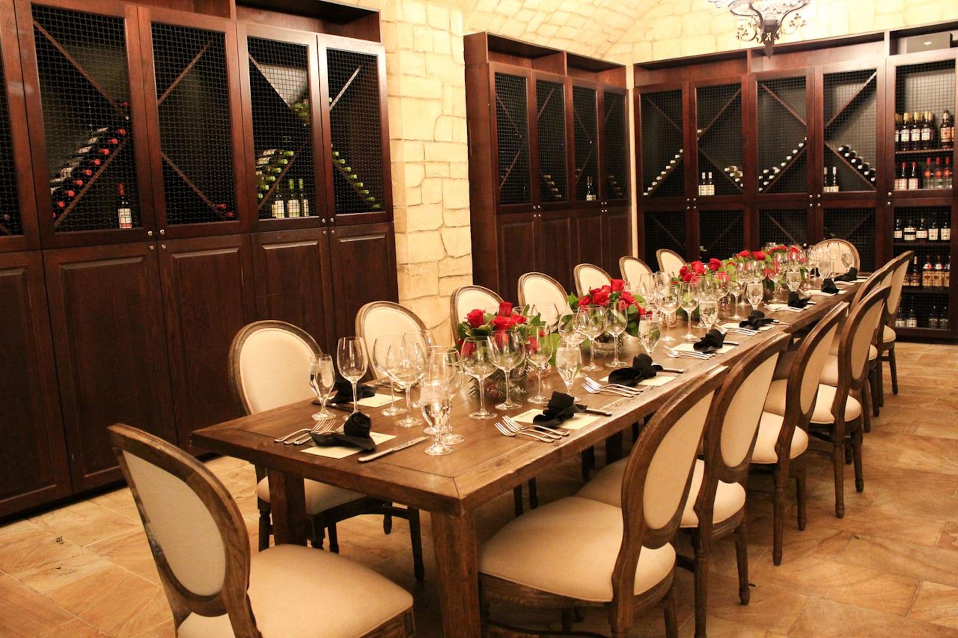A long dining table set for a formal meal, surrounded by wooden chairs, with wine racks in the background.