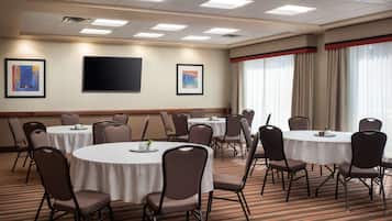 Un salón de conferencias con mesas redondas y sillas, un televisor de pantalla plana, obras de arte enmarcadas en las paredes y ventanas amplias con cortinas.