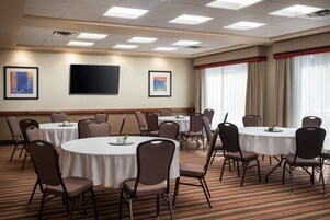 Un salón de conferencias con mesas redondas y sillas, un televisor de pantalla plana, obras de arte enmarcadas en las paredes y ventanas amplias con cortinas.
