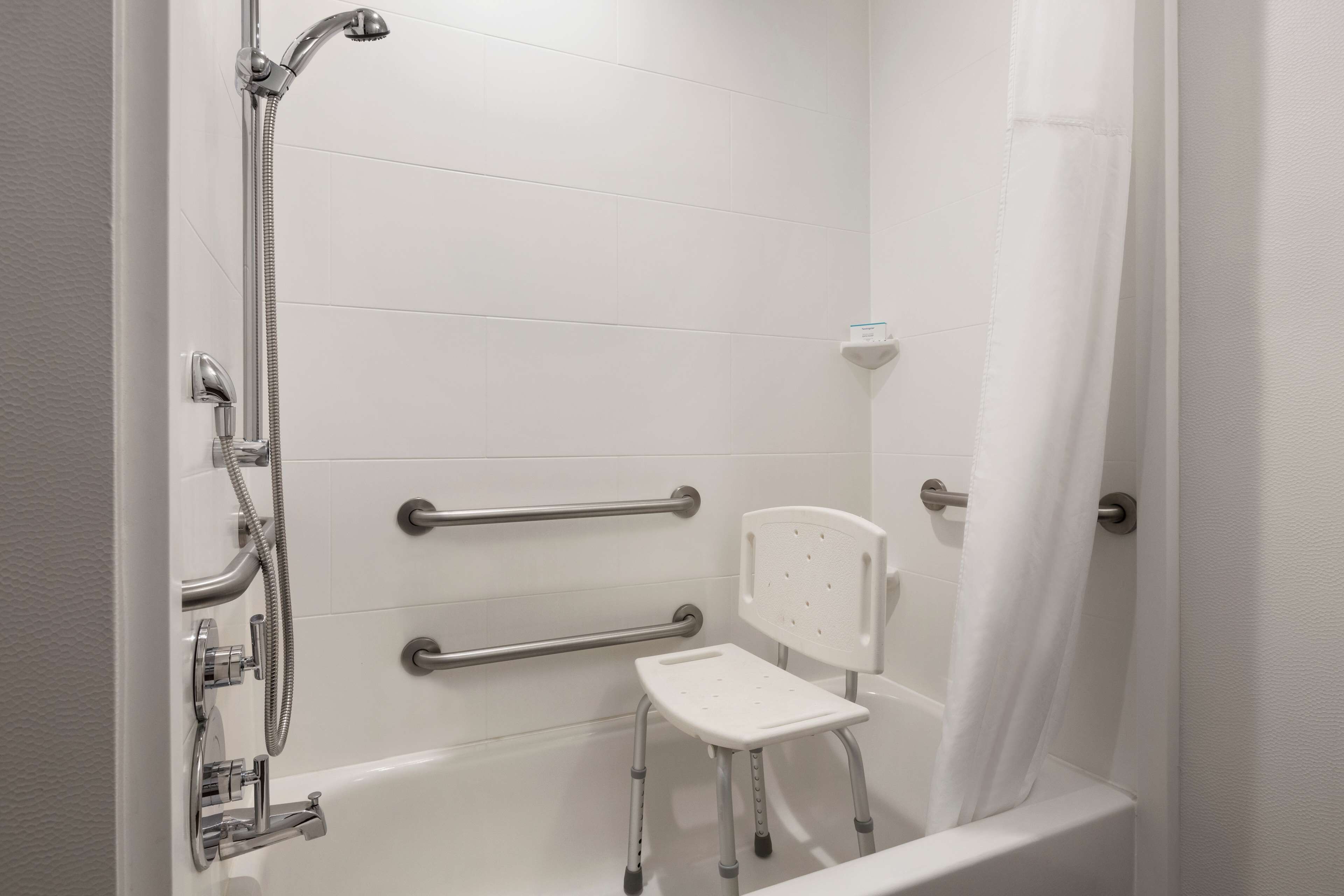 Un baño con una bañera blanca, una ducha con cabezal de ducha manual y una silla blanca con reposabrazos.