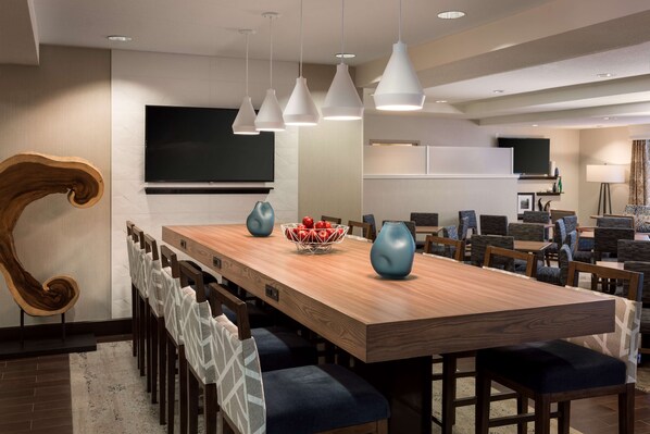 A modern dining area with a long wooden table, blue chairs, and pendant lights. - Hampton Inn-Denver West/Golden (Golden)