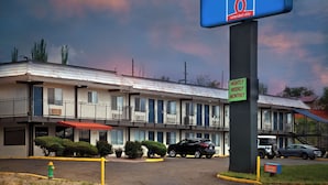 Ein Motel mit einem Schild für Studio 6 Extended Stay, das Tages-, Wochen- und Monatsangebote anzeigt. Es gibt einen Parkplatz mit mehreren Autos und einen klaren Himmel.