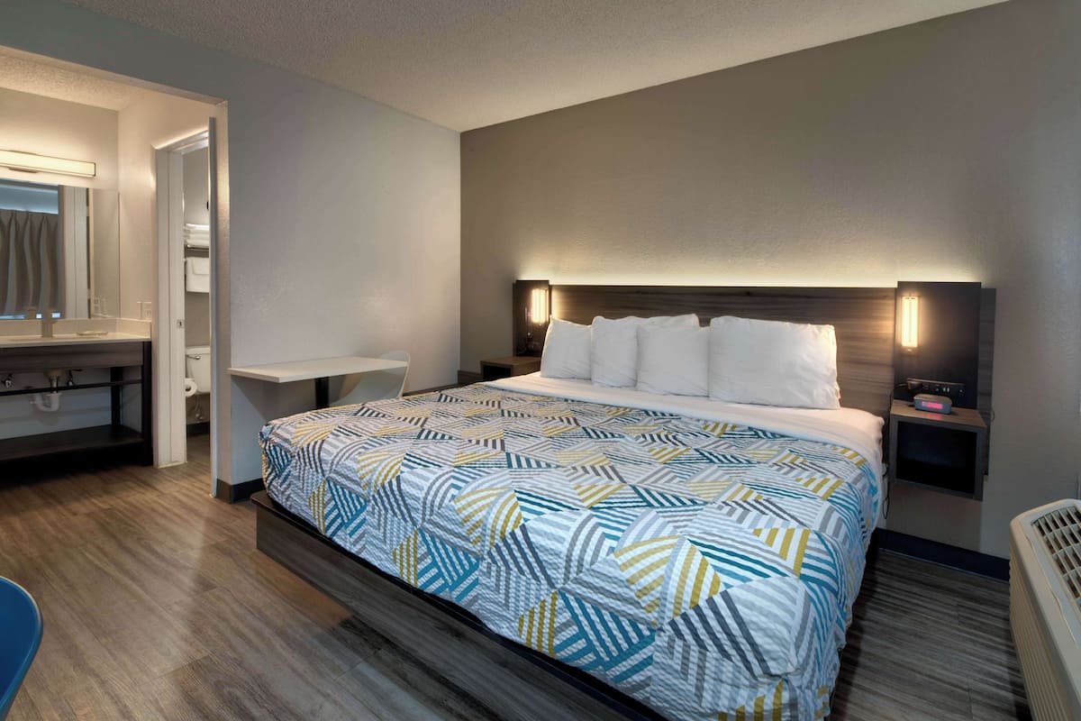 A hotel room with a large bed, a bedside table, a nightstand, a mirror, and a bathroom visible through an open door.