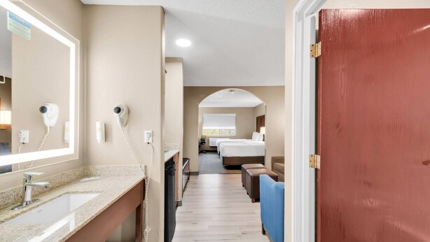 Un pasillo de hotel con un baño a la izquierda, una zona de camas en el centro y una puerta roja a la derecha.