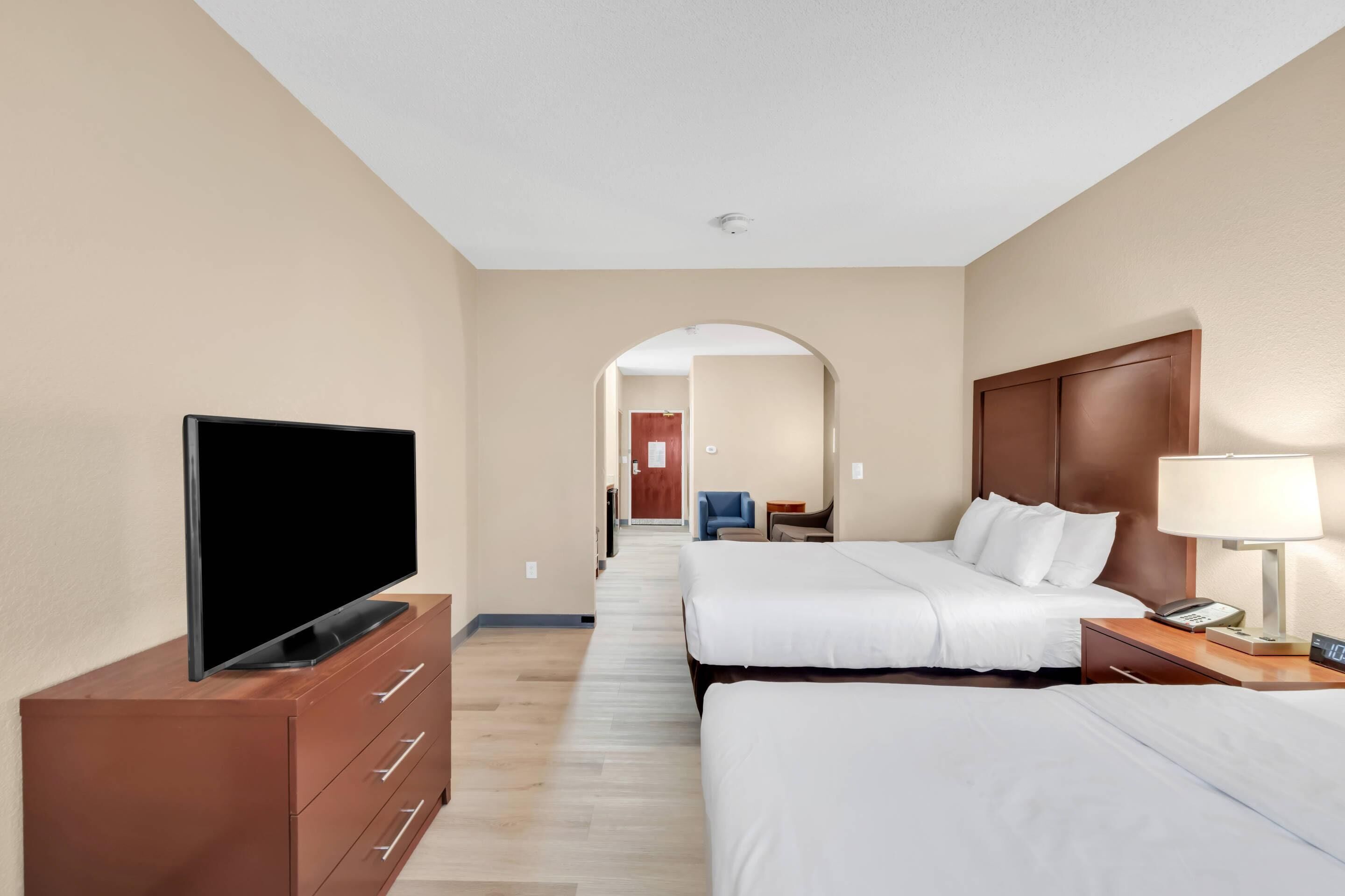 Habitación de hotel con televisor sobre un tocador de madera, dos camas con ropa blanca, una mesita de noche con lámpara y un pasillo que conduce a otra habitación.