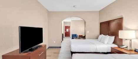 Habitación de hotel con televisor sobre un tocador de madera, dos camas con ropa blanca, una mesita de noche con lámpara y un pasillo que conduce a otra habitación.