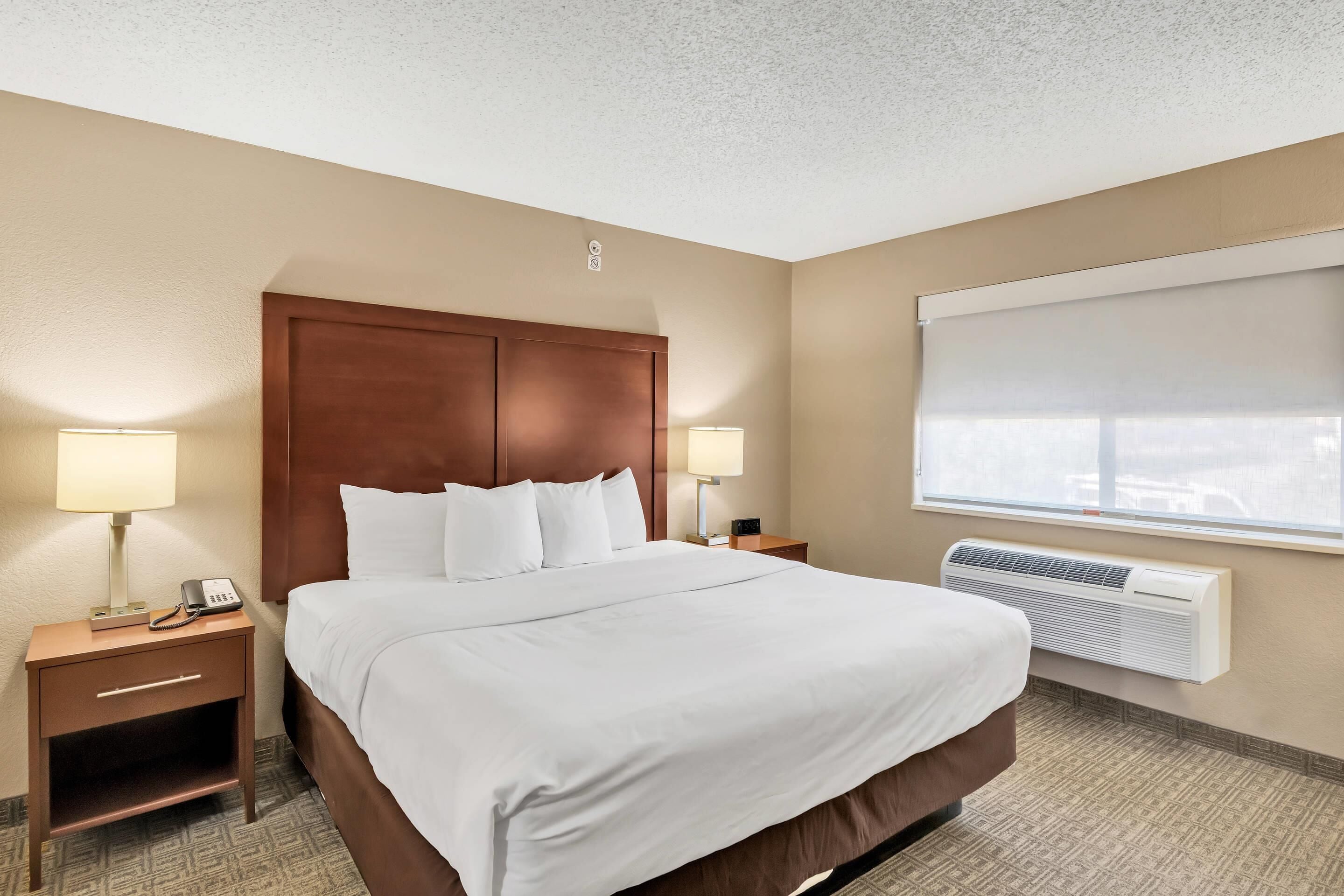 A hotel room with a large bed, two bedside tables with lamps, a wall-mounted air conditioner, and a window with a blind.