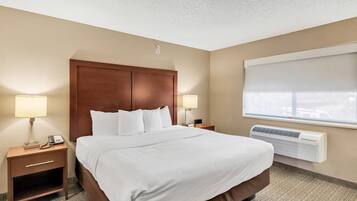 A hotel room with a large bed, two bedside tables with lamps, a wall-mounted air conditioner, and a window with a blind.