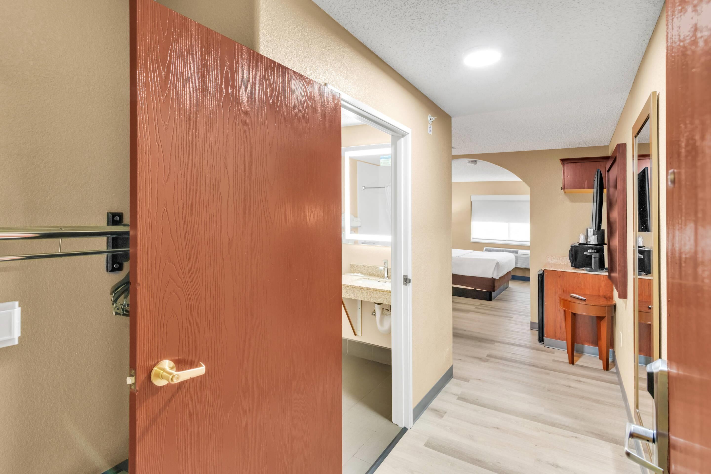 A hotel hallway with a wooden door, a bed, a desk, and a small table.