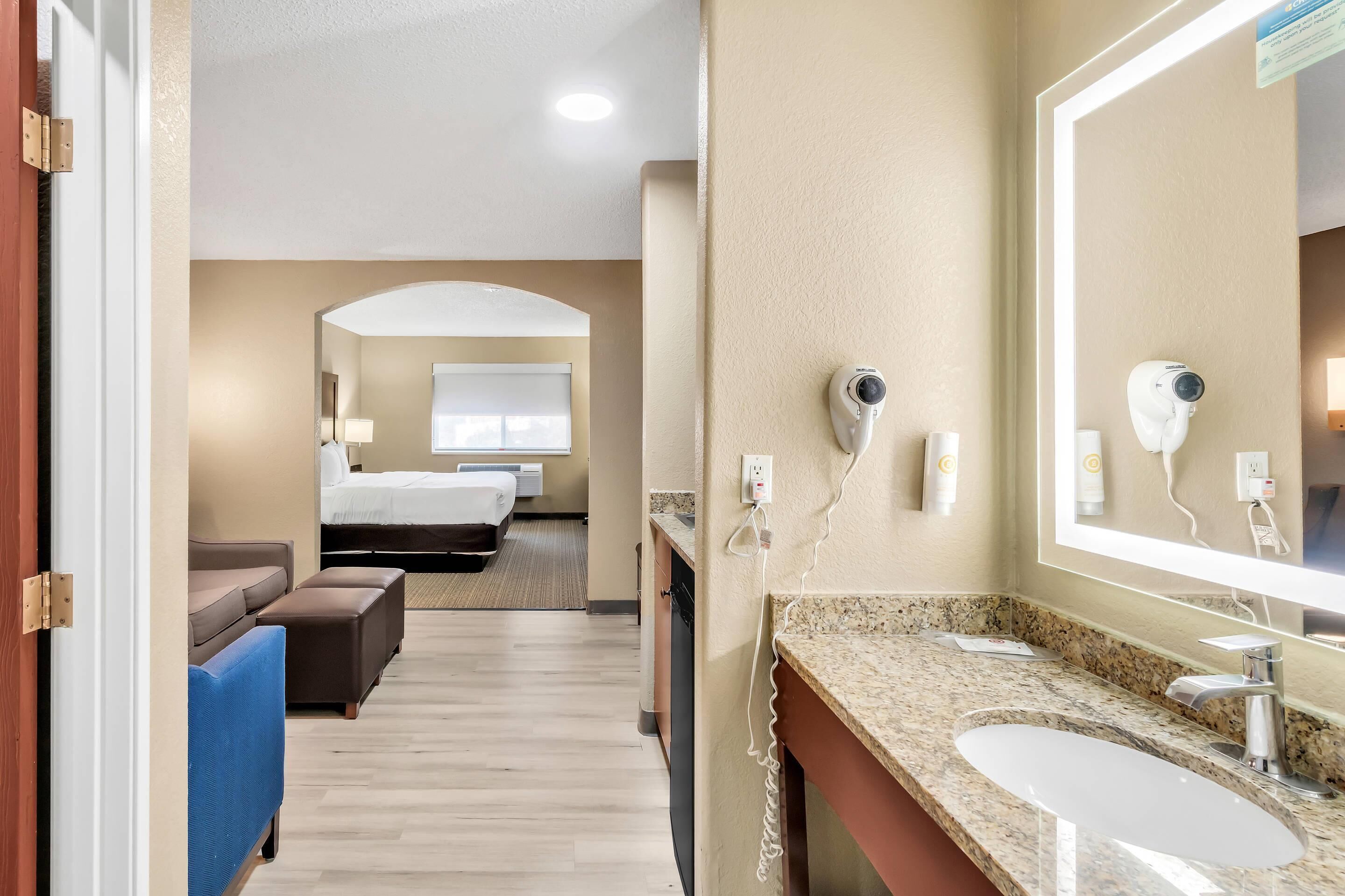 A hotel room with a bed, a sofa, a small table, a bathroom with a sink and mirror, and a security camera.