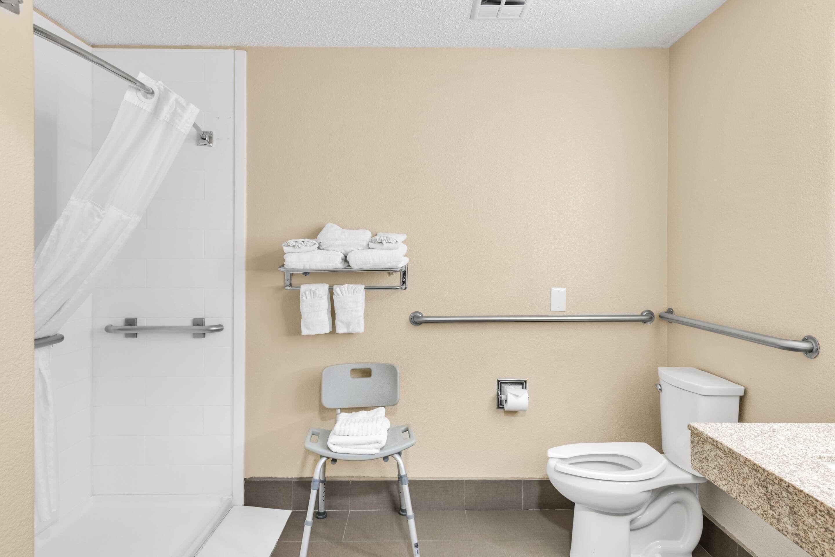 A bathroom with a shower, a toilet, a grab bar, a chair, and a towel rack.