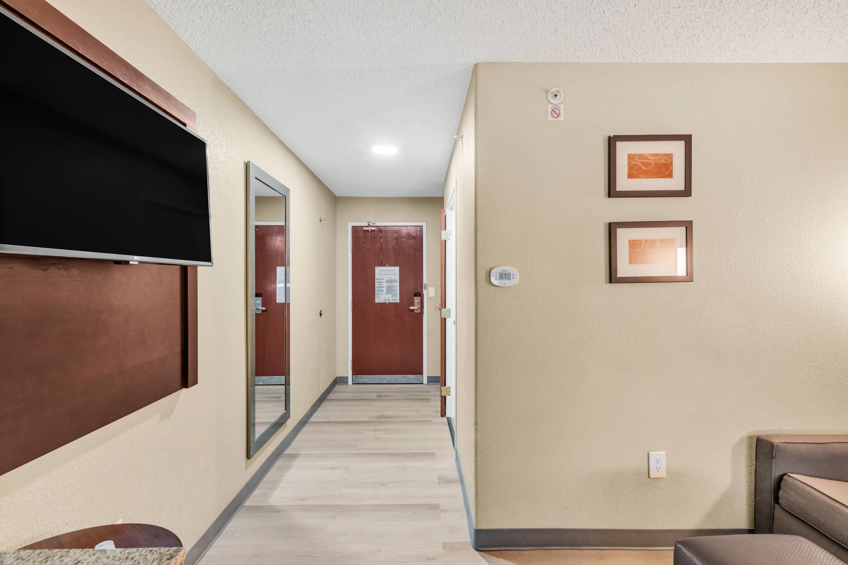 A hotel hallway with a flat-screen TV mounted on the wall, a wooden door at the end, and two framed pictures on the wall.