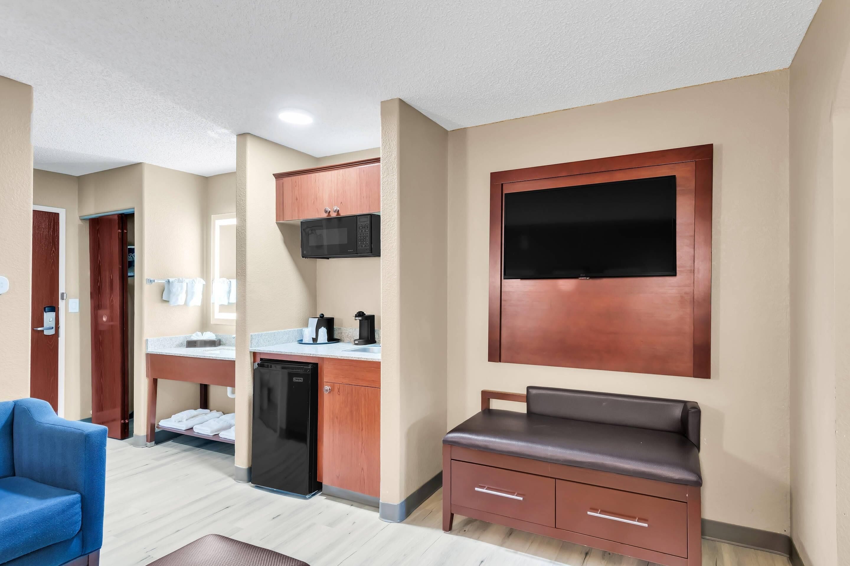Habitación de hotel con kitchenette, zona de estar con sofá y una mesita, y un televisor de pantalla plana grande montado en la pared.