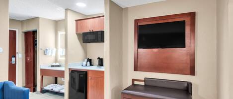 Habitación de hotel con kitchenette, zona de estar con sofá y una mesita, y un televisor de pantalla plana grande montado en la pared.