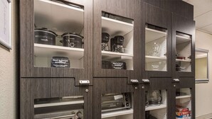 A kitchen with built-in appliances including a toaster oven, a slow cooker, and a coffee maker. There are shelves with storage containers and a sign asking if someone needs to borrow something.