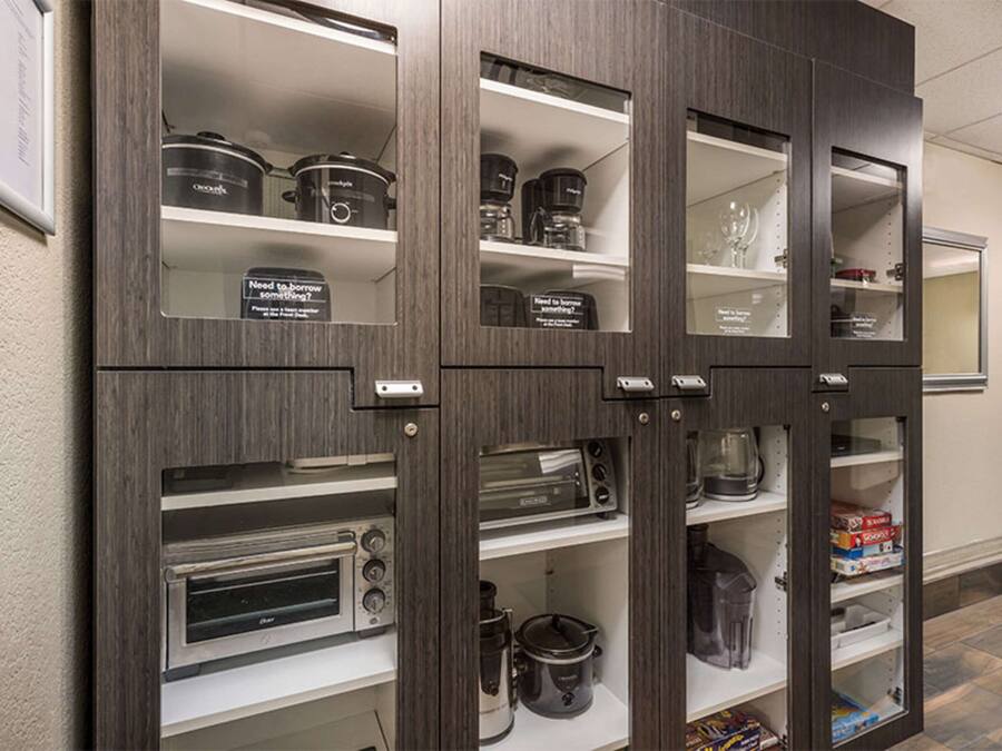 A kitchen with built-in appliances including a toaster oven, a slow cooker, and a coffee maker. There are shelves with storage containers and a sign asking if someone needs to borrow something.