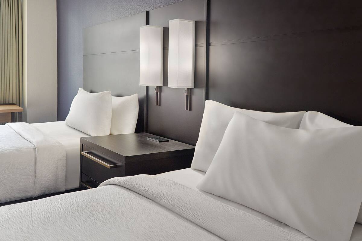 Une chambre d’hôtel avec deux lits, une tête de lit sombre et deux appliques murales.