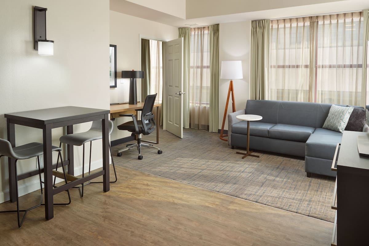 Une chambre d’hôtel moderne avec un plancher en bois, un canapé gris, un bureau avec deux chaises, une lampe sur pied et une grande fenêtre avec des rideaux.