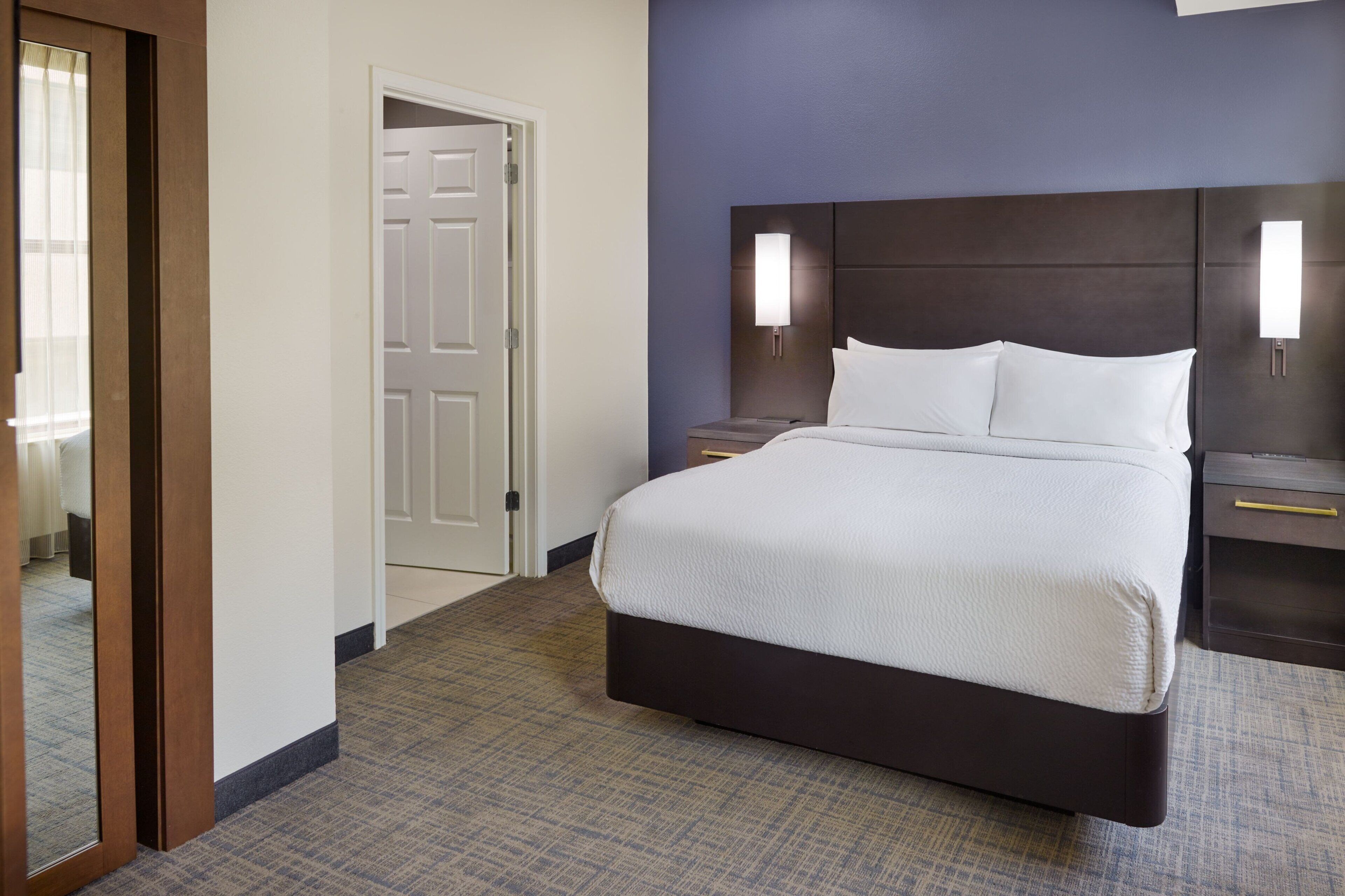 Quarto de hotel com uma cama grande, uma mesa de cabeceira, uma luminária na parede e uma porta que dá para outro quarto.
