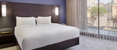 A hotel room with a large bed, a dark headboard, two bedside lamps, and a view of the outdoors through large windows.