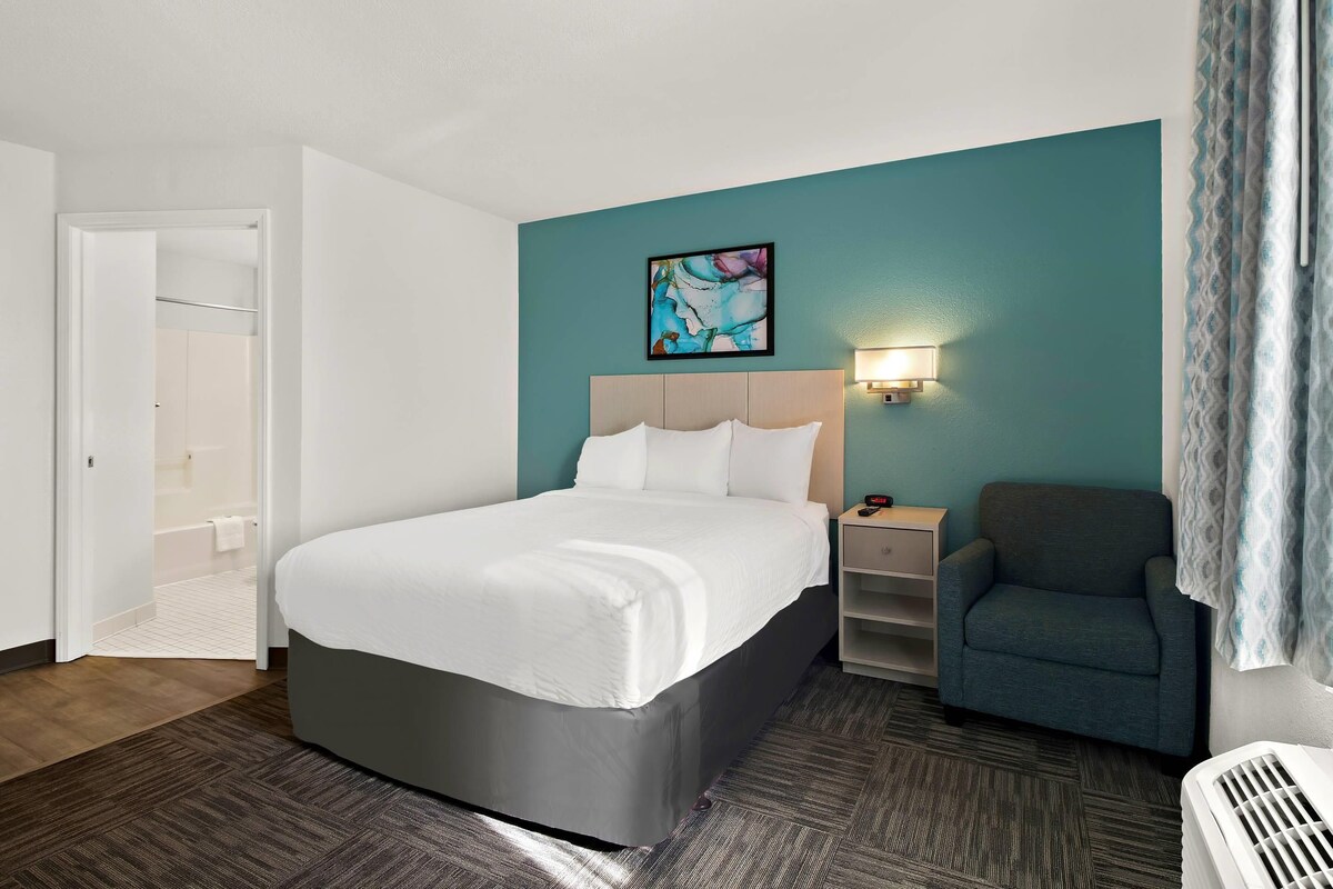 A hotel room with a bed, a chair, a nightstand, a wall-mounted lamp, and a door leading to a bathroom.